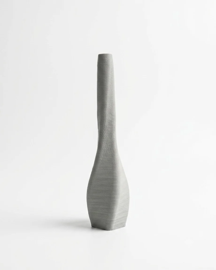 Vase en céramique gris imprimé en 3D, au long col étroit et à la base facettée, présenté sur fond clair.